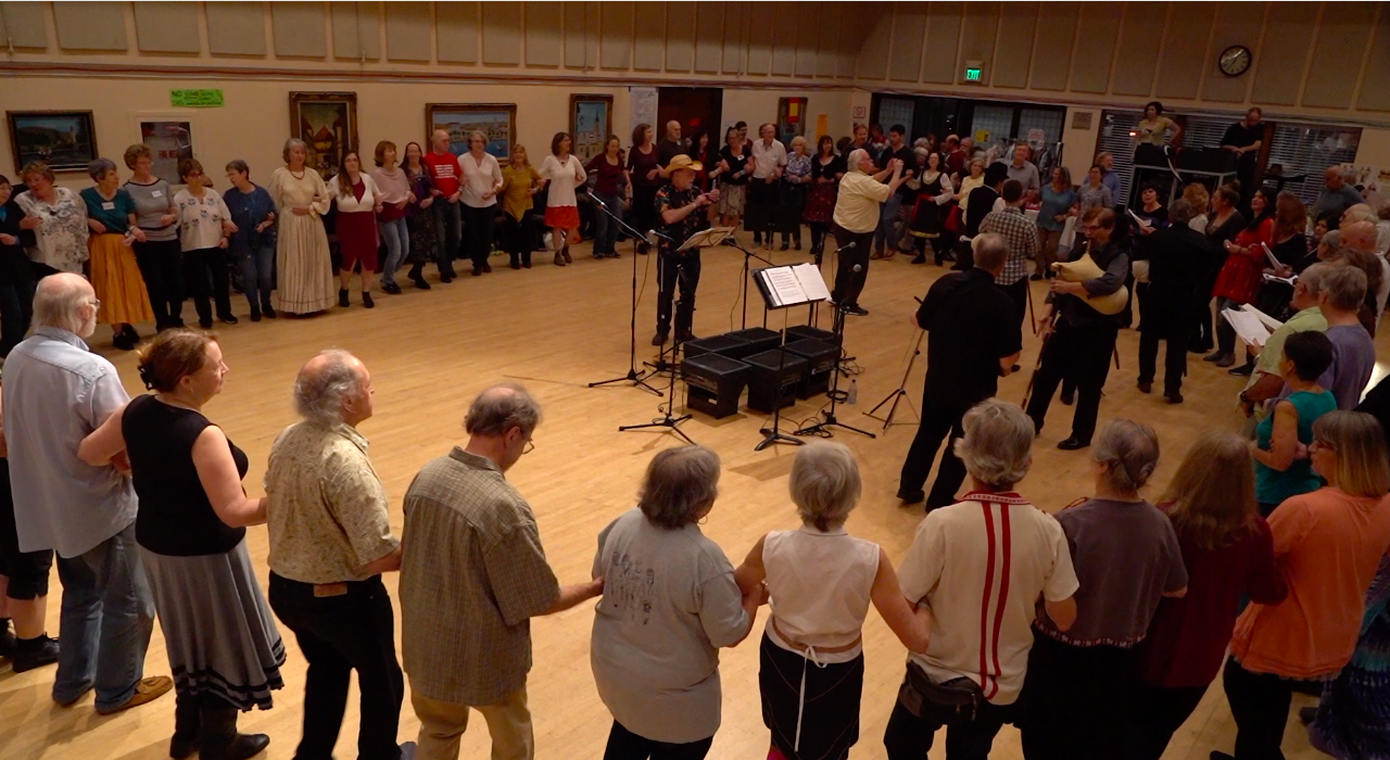 Dancers linked arm-in-arm in a large circle, with musicians playing in the center