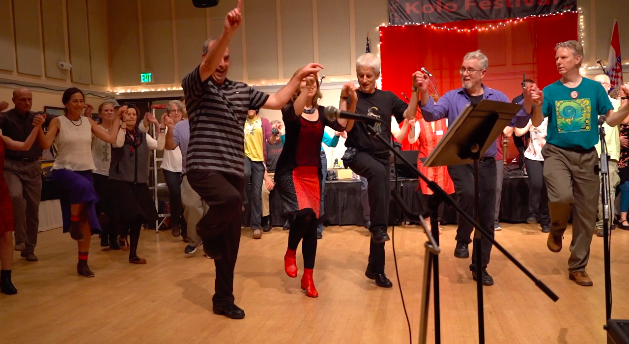 Dancers mid-step with hands joined and one leg raised, led by the dance instructor