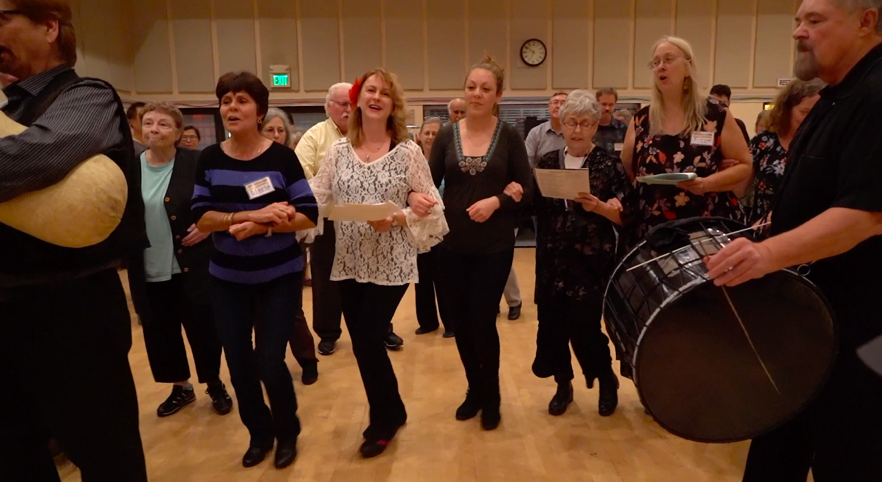 Four women singing from sheet music with joined hands, accompanied by a large drum