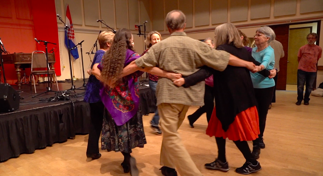 Nine dancers in a circle with arms around each other's backs