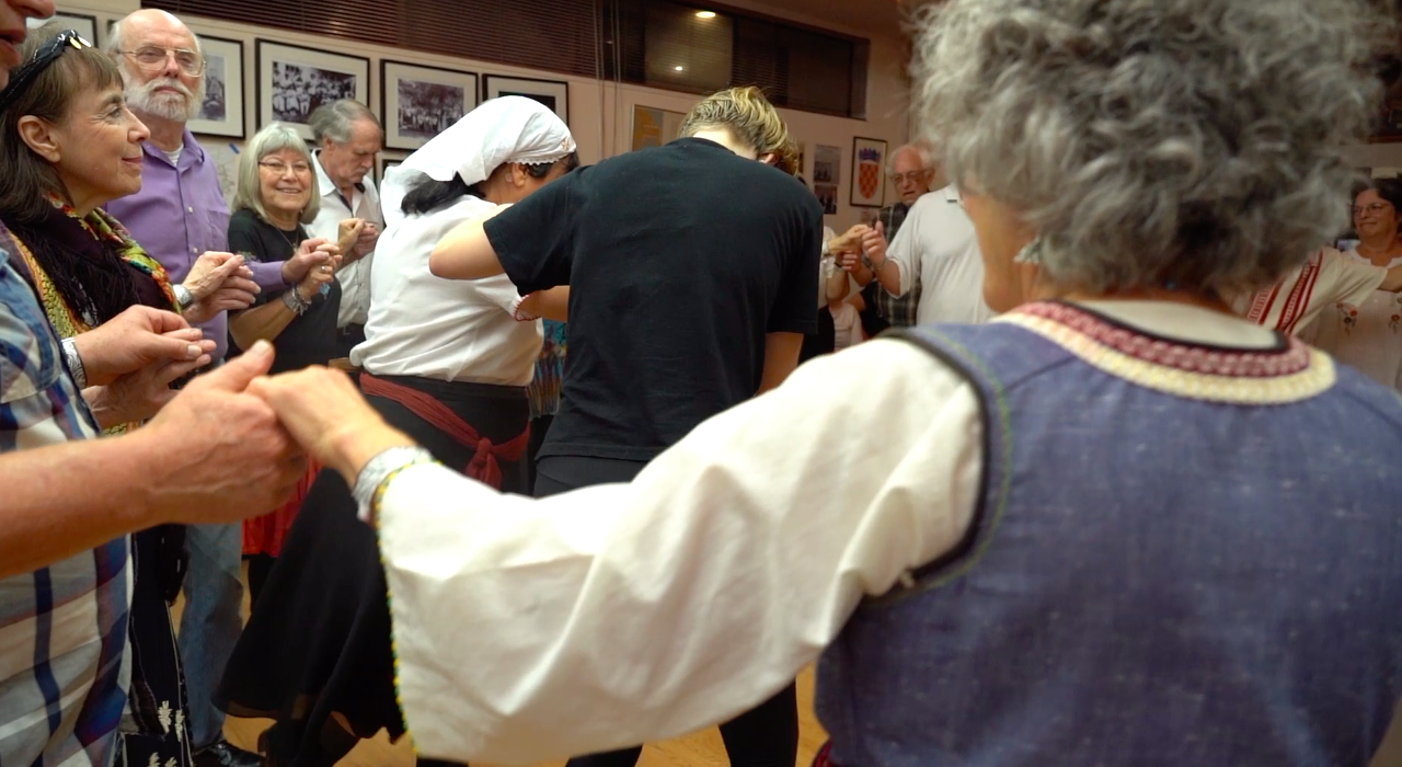 Festival attendees dancing in a circle with joined hands, one being shown a step